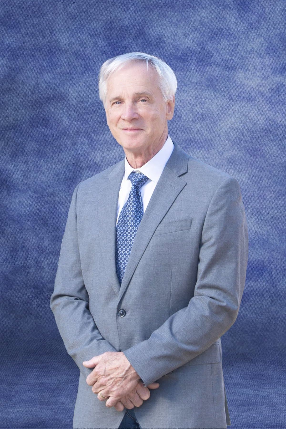 John Armbruster, in a grey suit and blue tie, photographed against a blue studio backdrop.